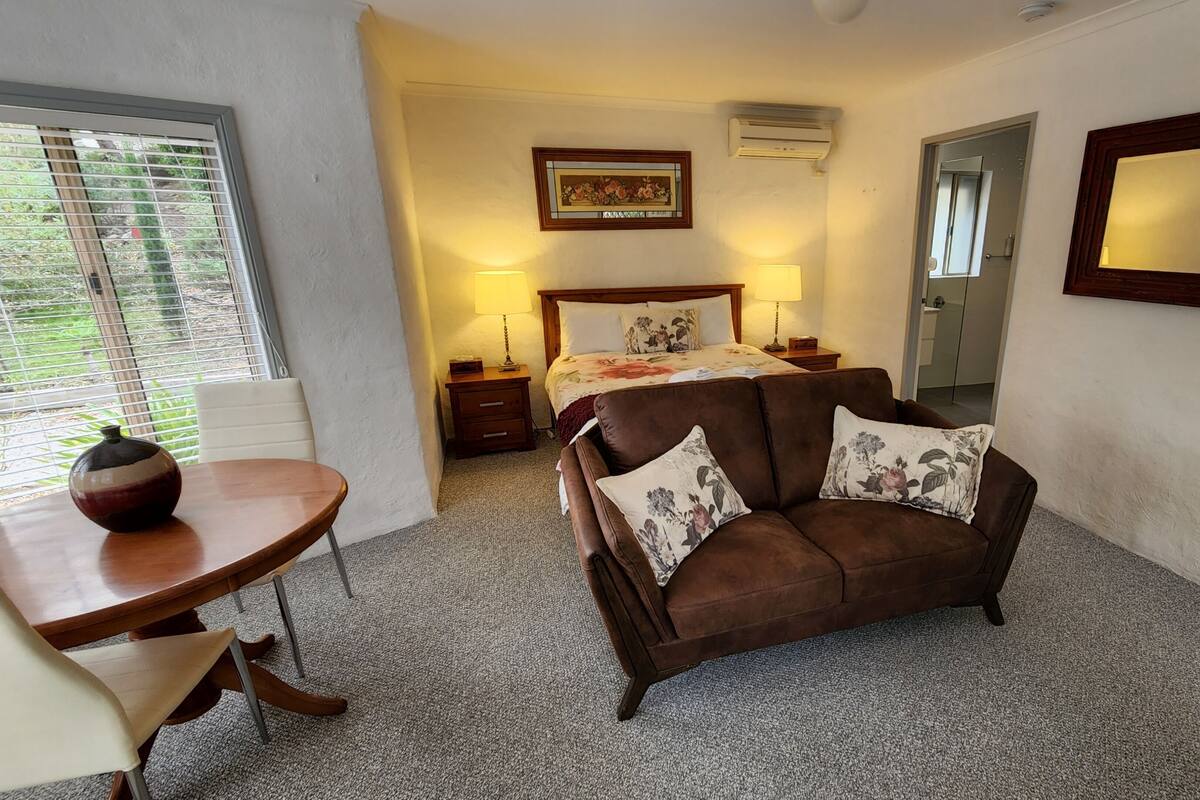 Une chambre à coucher avec un lit, un canapé marron agrémenté de coussins à motifs floraux, une table de chevet avec un vase, et un grand miroir accroché au mur.
