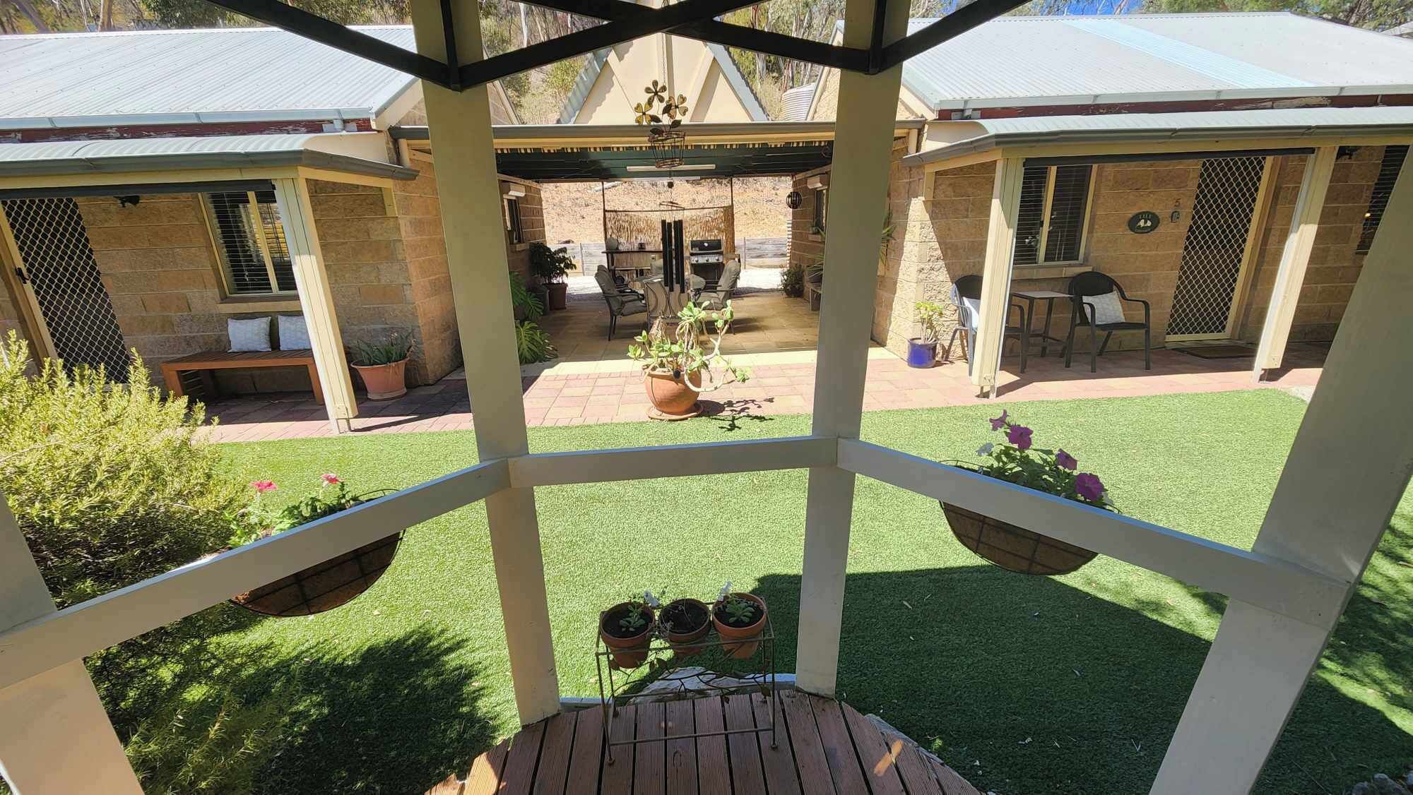 Ein überdachter Außenbereich mit Terrasse, Blumentöpfen und Blick auf einen Garten mit Grill und Sitzgelegenheit.