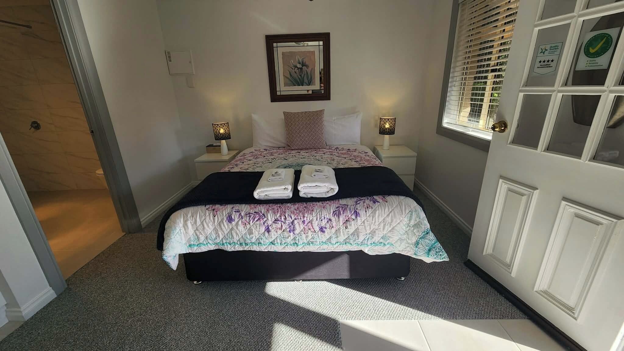 A bedroom with a bed, two bedside lamps, a framed picture on the wall, and a window with blinds.