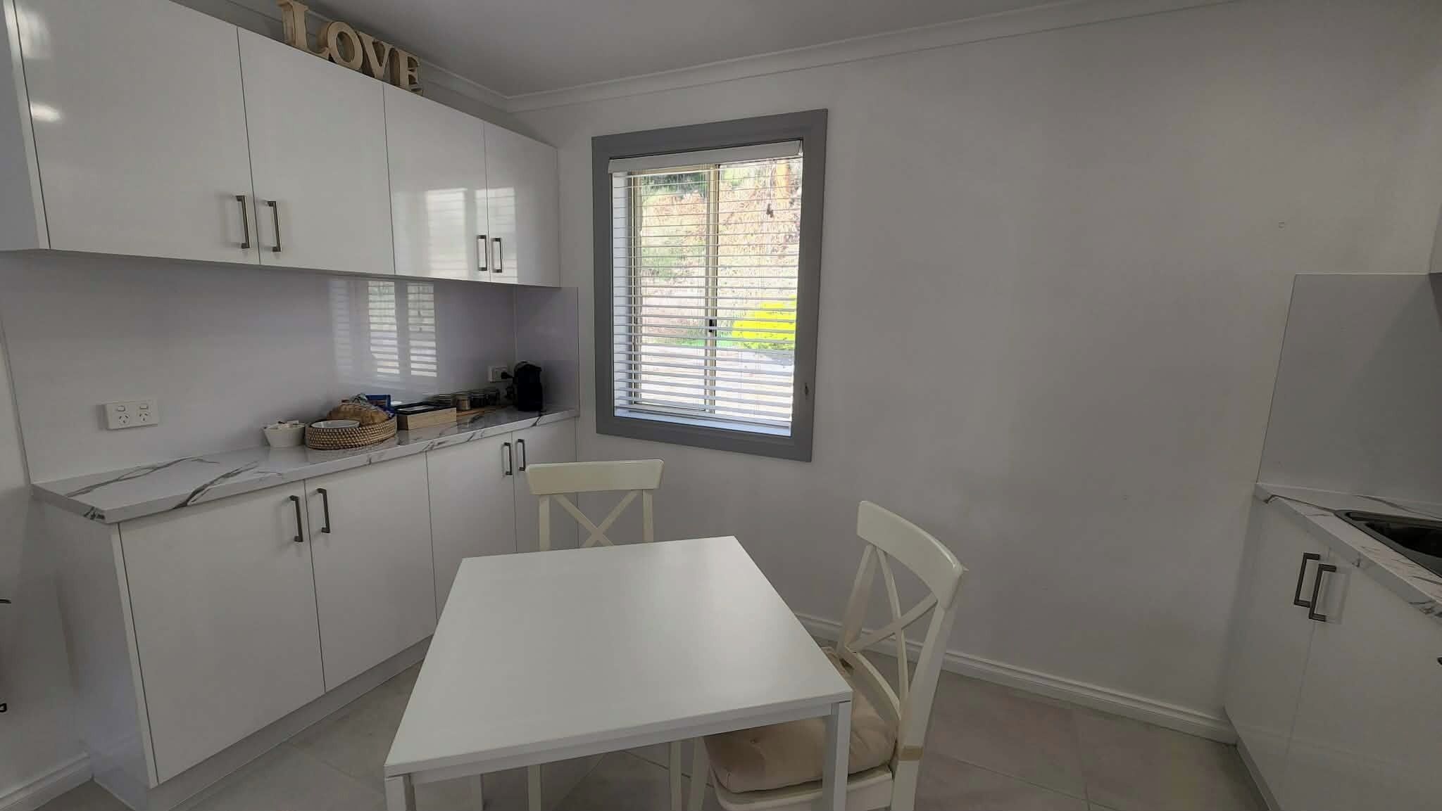 Una cocina pequeña con armarios blancos, una ventana con persianas, una mesa blanca y una silla.