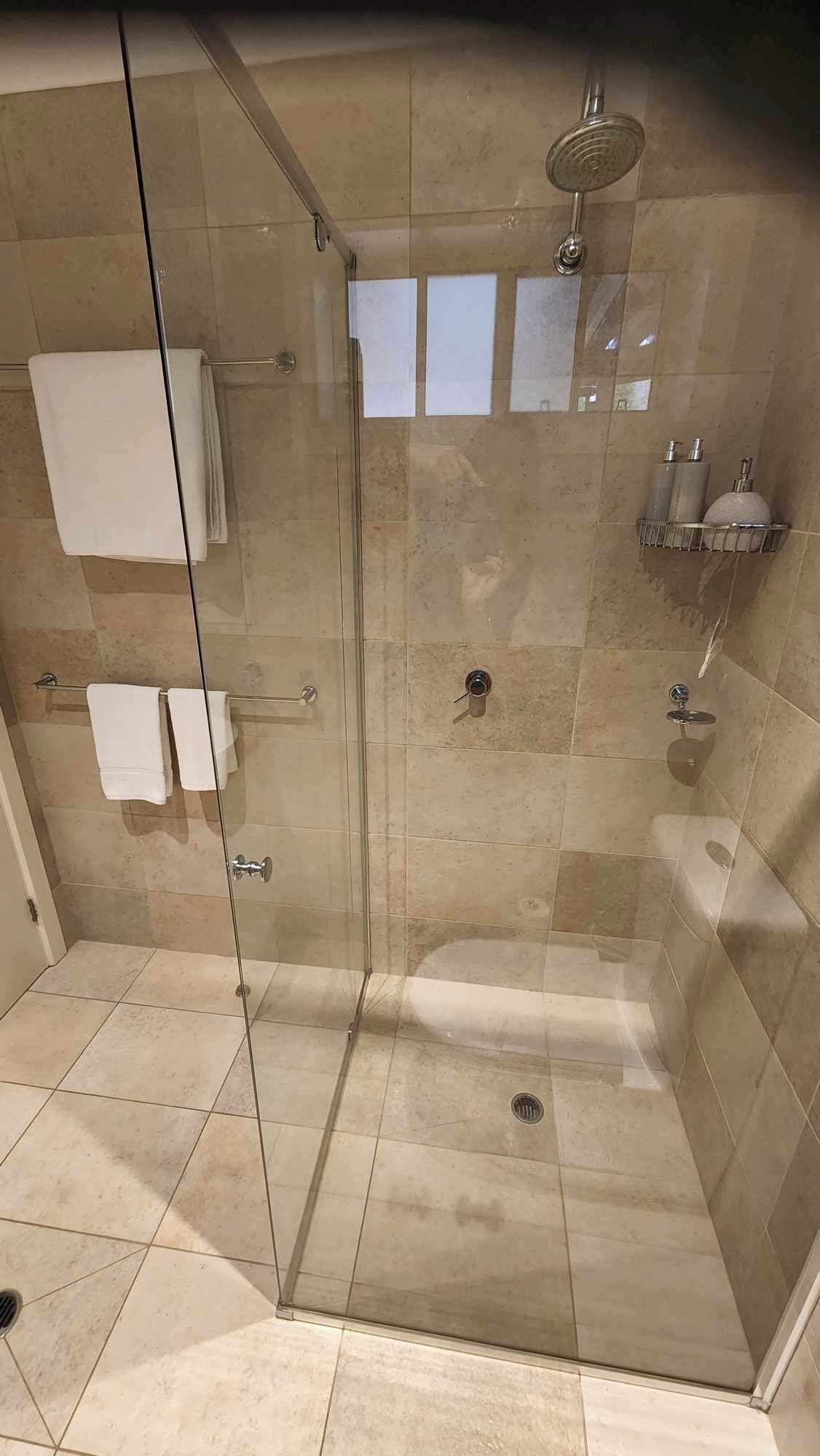 A glass-enclosed shower stall with a showerhead, a towel rack with folded towels, and a small shelf with toiletries.