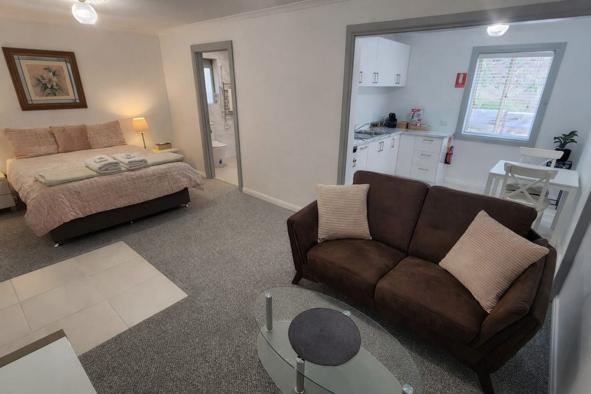 Une chambre à coucher avec un lit, un canapé marron, une petite table et un coin cuisine visible à travers une porte ouverte.