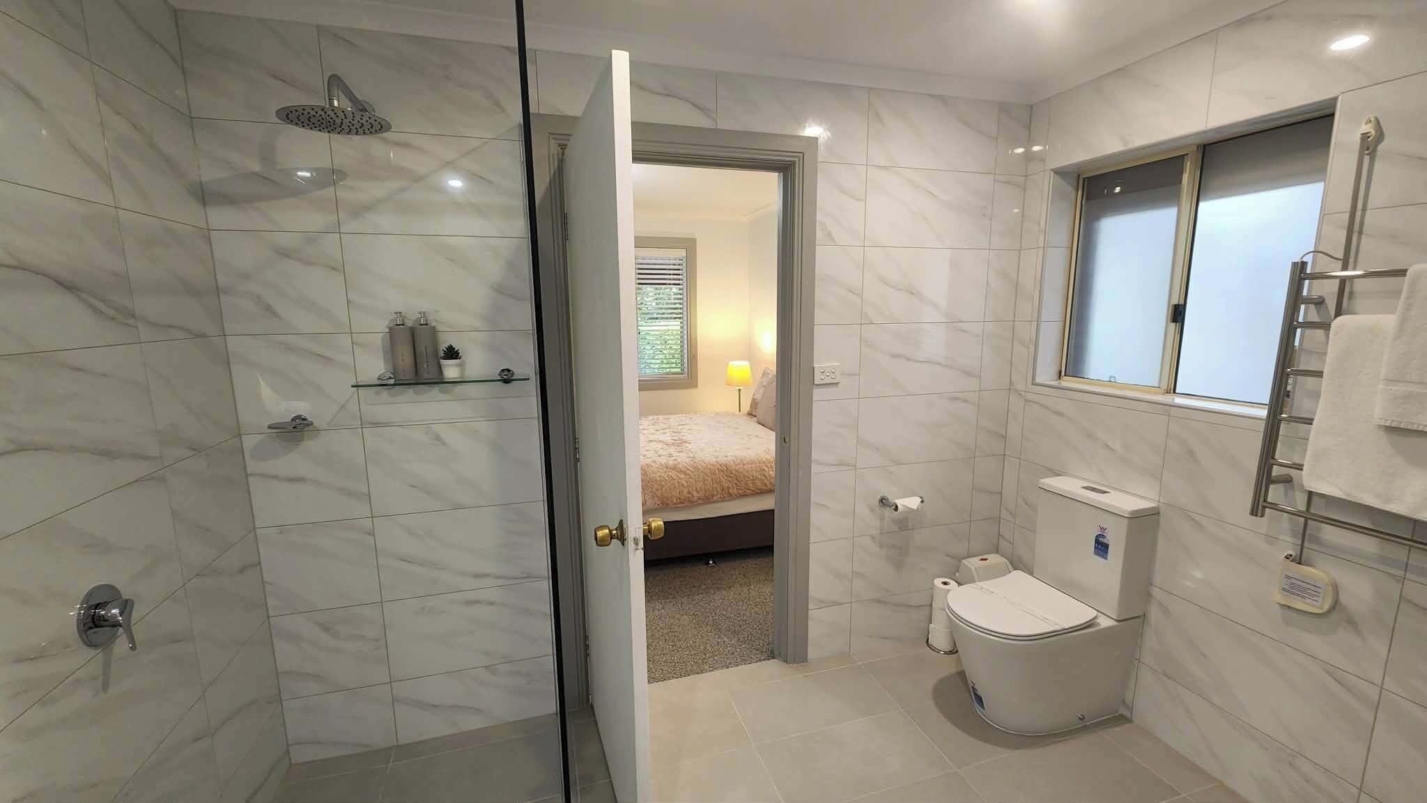 A bathroom with a shower, toilet, and a bed visible through an open door.