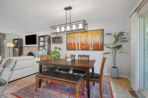 Uma área de refeições com mesa de madeira, bancos e cadeiras. Há um quadro grande na parede, um vaso de planta e uma TV de tela plana instalada acima da lareira.