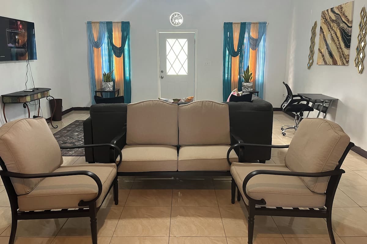A living room with a sofa, two armchairs, a TV, a clock, and a door with curtains.