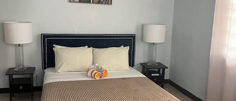 A neatly made bed with a striped bedspread, two bedside tables with lamps, a bench at the foot of the bed, and a floral artwork on the wall.