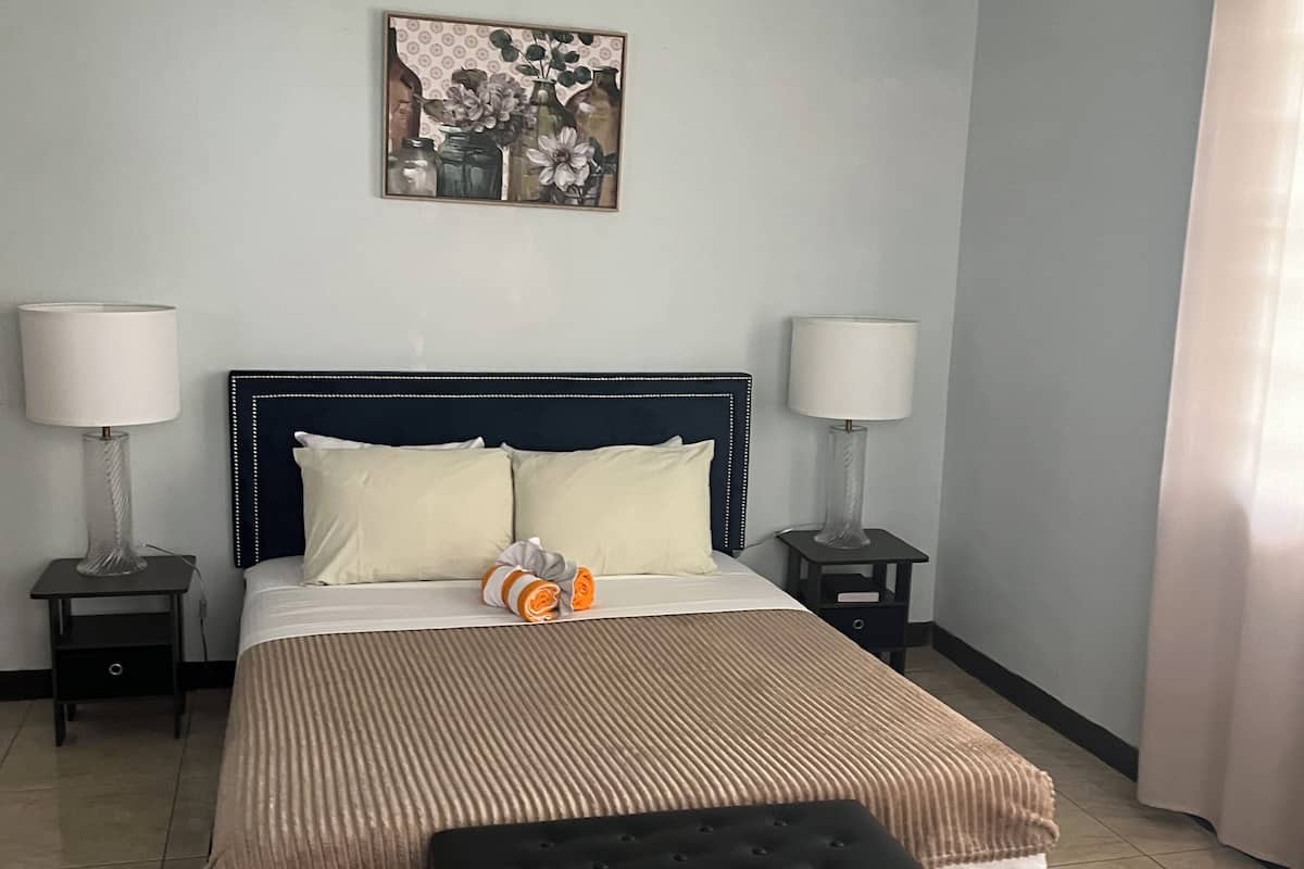 A neatly made bed with a striped bedspread, two bedside tables with lamps, a bench at the foot of the bed, and a floral artwork on the wall.