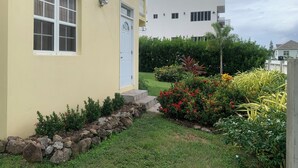 Il s’agit d’un immeuble d’habitation à la façade jaune, aux fenêtres blanches et à la porte blanche. Un jardin, où l’on peut admirer diverses plantes et fleurs, se trouve devant l’immeuble.