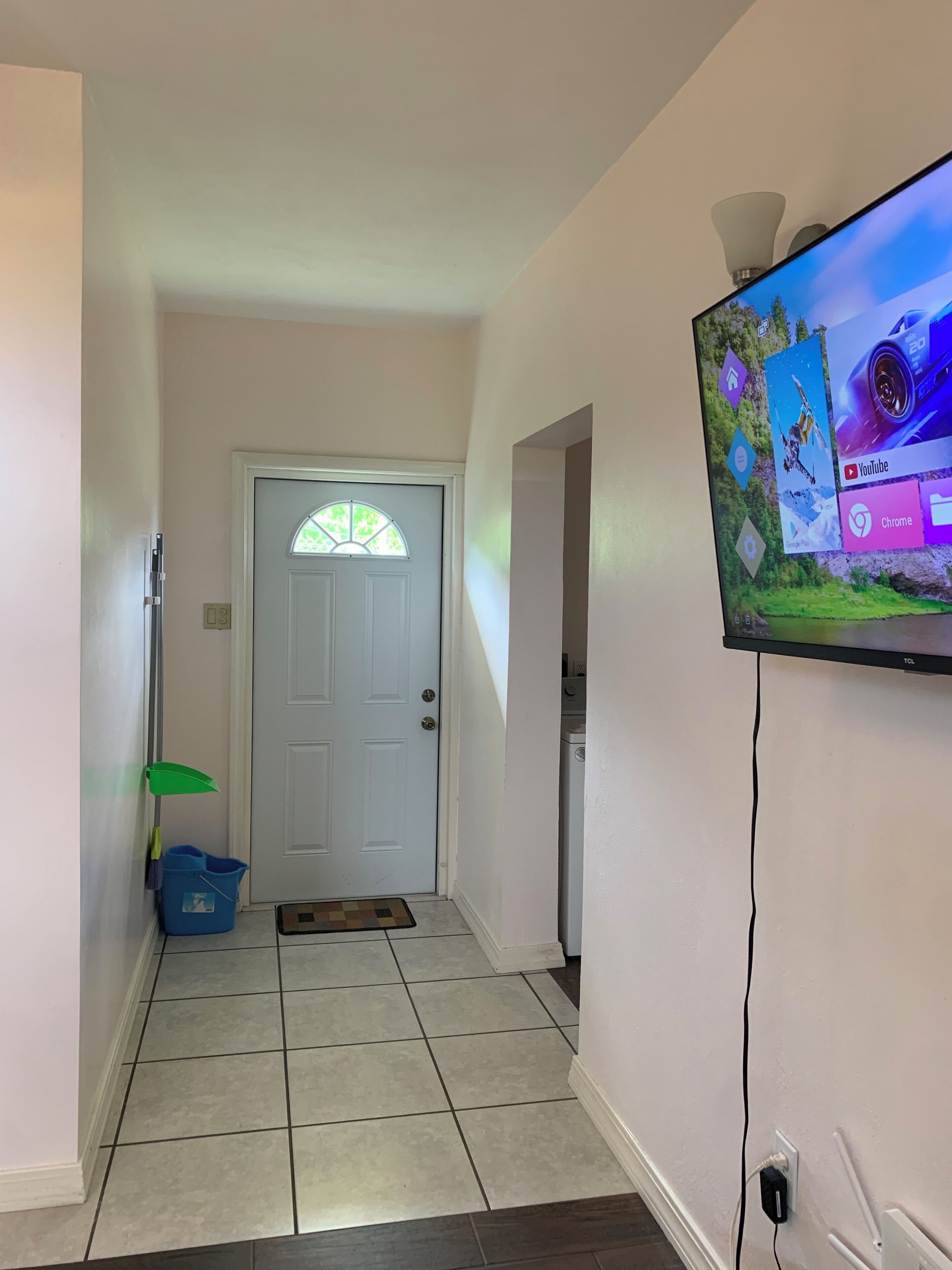 A hallway with a tiled floor, a white door with a window, a blue storage bin, and a wall-mounted television displaying various icons.