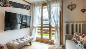 Una sala moderna con un televisor de pantalla plana grande, un banco acolchado, un sofá con cojines estampados y vistas a una terraza de madera y follaje exterior.