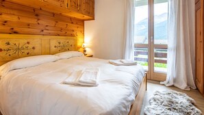 Un dormitorio con cabecera de madera, cama con sábanas blancas, mesita de noche con lámpara y vistas a montañas a través de una ventana.