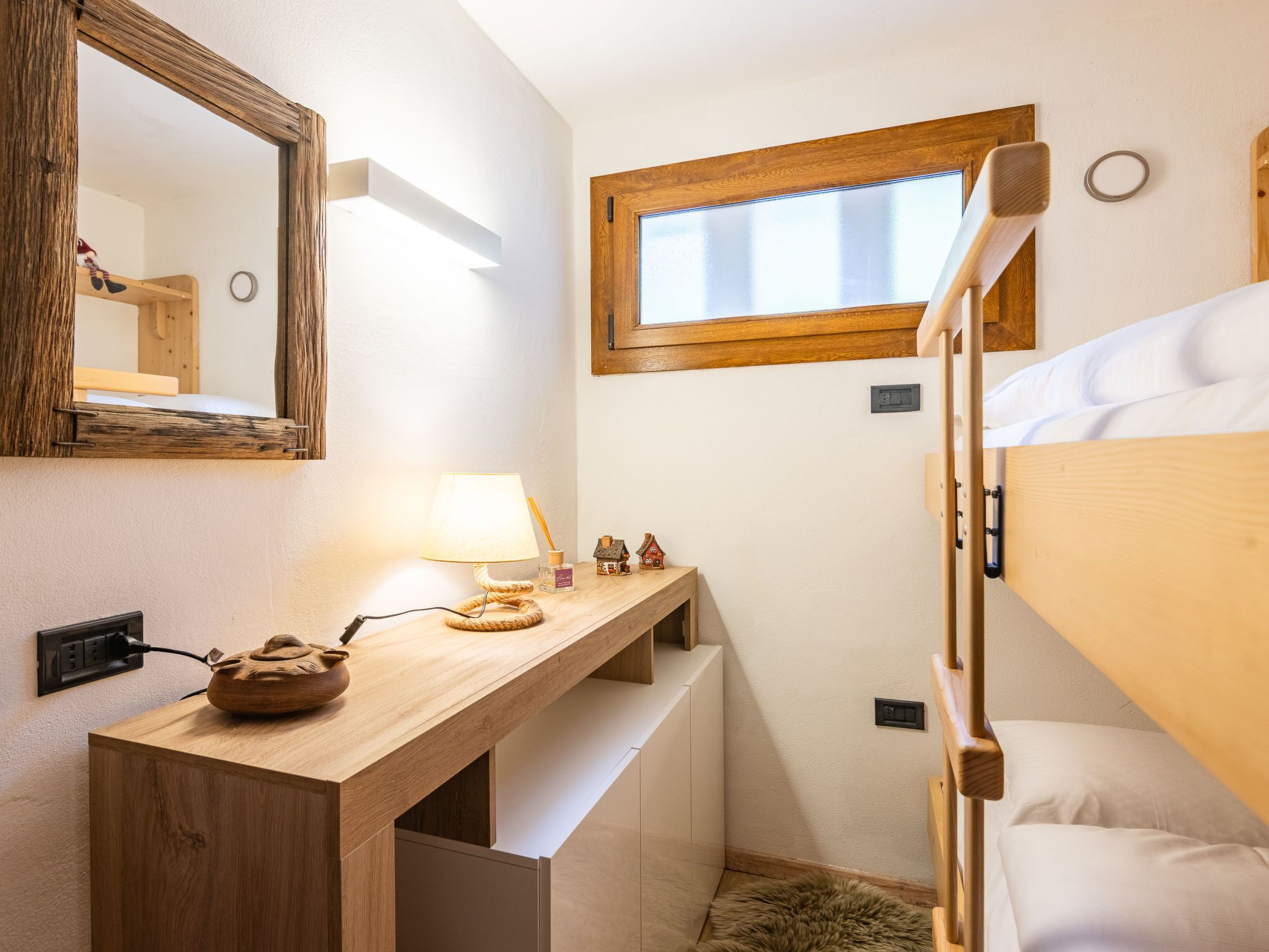 Een compacte slaapkamer met een stapelbed, een houten bureau met lamp, een klein plankje met decoratieve spullen, en een raam met zonwering.