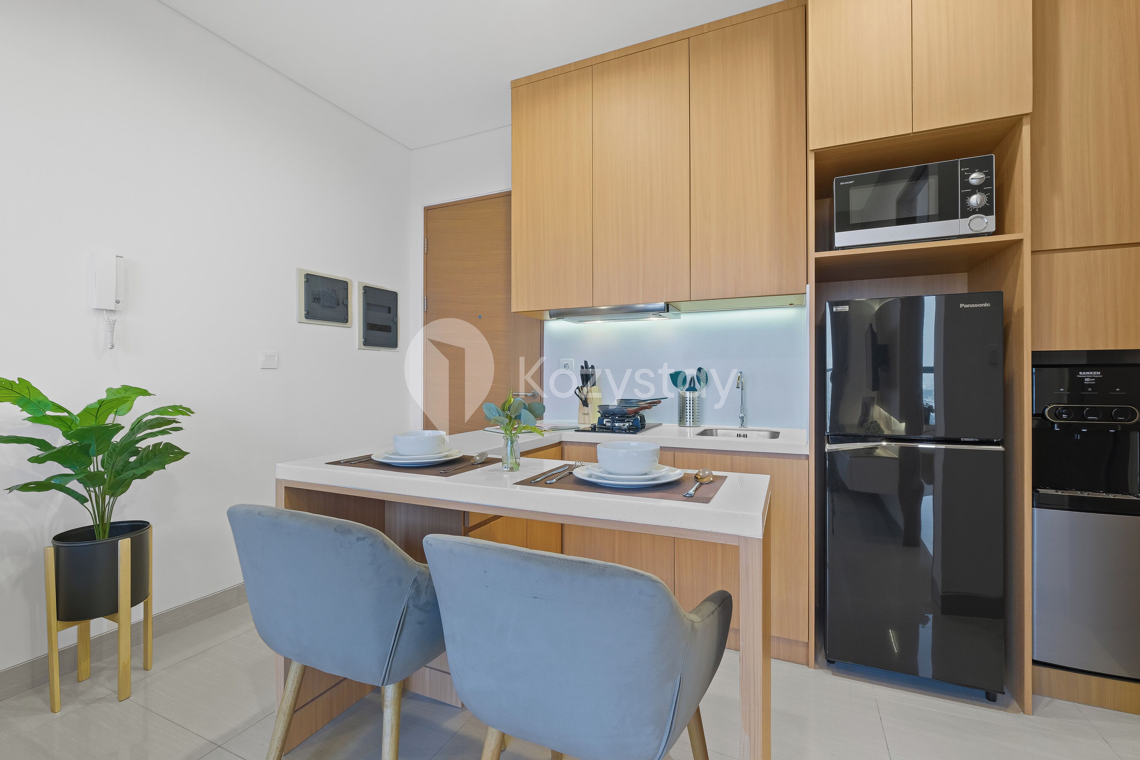 Una cocina moderna con armarios de madera, un microondas integrado, un refrigerador de acero inoxidable y un comedor con dos sillas y una pequeña planta en maceta.