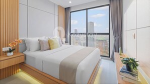 A modern hotel room with a large bed, a wooden nightstand, a bedside table with a plant, and a view of the city through large windows.