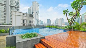 A rooftop pool with a wooden deck, surrounded by greenery and city buildings.