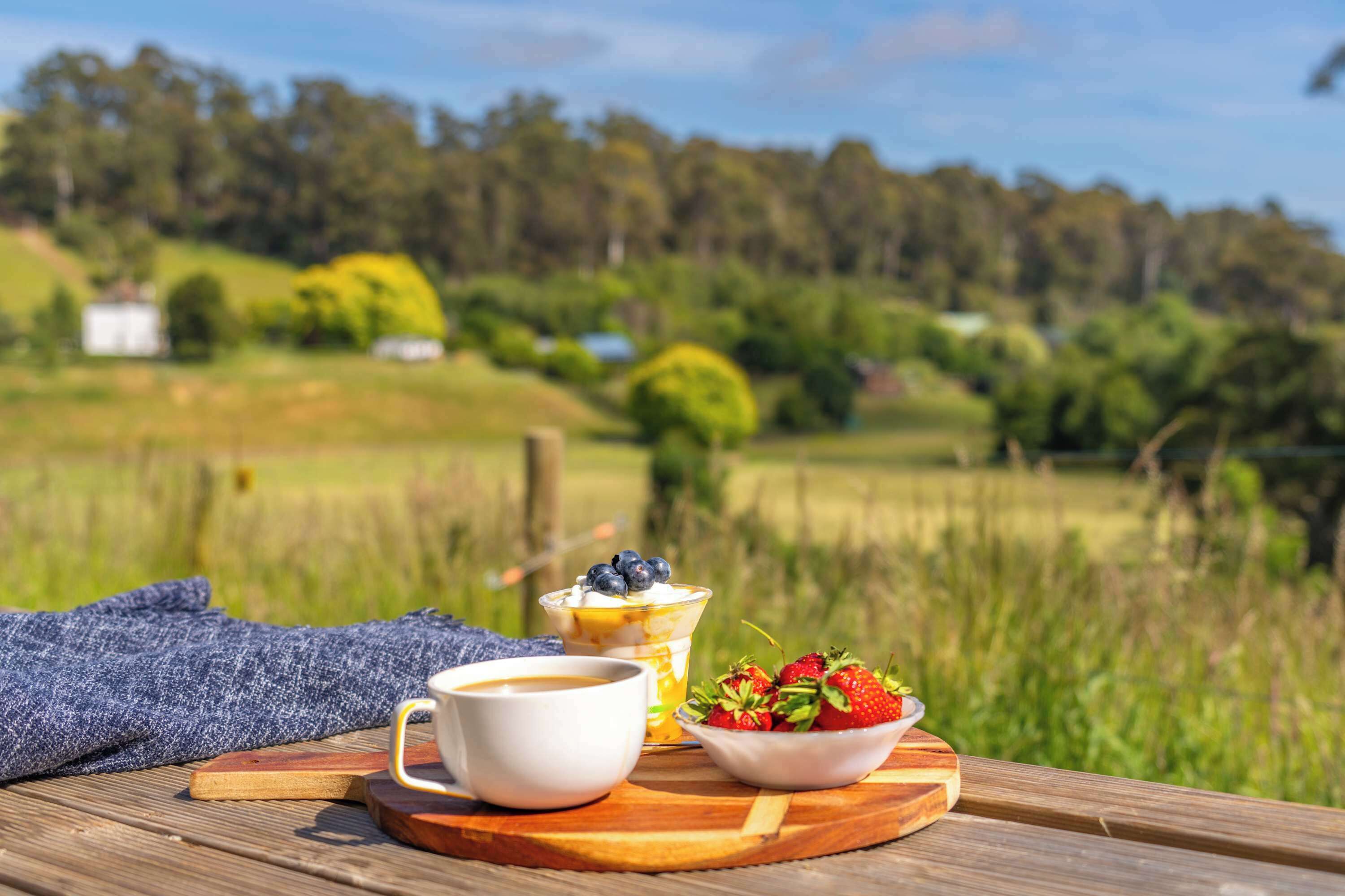 A wooden table with a cup of coffee, a bowl of strawberries, and a dessert with blueberries and whipped cream.