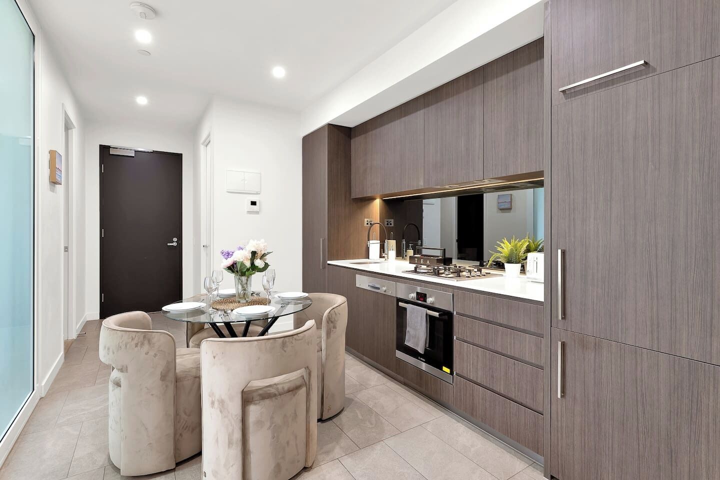 A modern kitchen with a dining area, featuring a glass table, cushioned chairs, and a built-in oven.