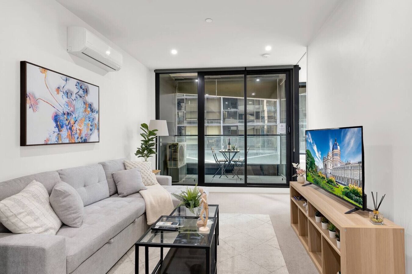 A modern living room with a grey sofa, a glass coffee table, a TV on a wooden stand, and a large window with a view of a building.