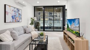 A modern living room with a grey sofa, a glass coffee table, a TV on a wooden stand, and a large window with a view of a building. - Central CBD Location | Pool & Gym | 2BR 2Bath (Melbourne)