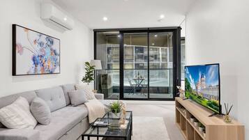 A modern living room with a grey sofa, a glass coffee table, a TV on a wooden stand, and a large window with a view of a building.