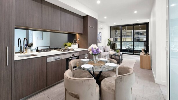 A modern kitchen with dark wood cabinets, a white countertop, and a dining area with a glass table and cushioned chairs. - Central CBD Location | Pool & Gym | 2BR 2Bath (Melbourne)