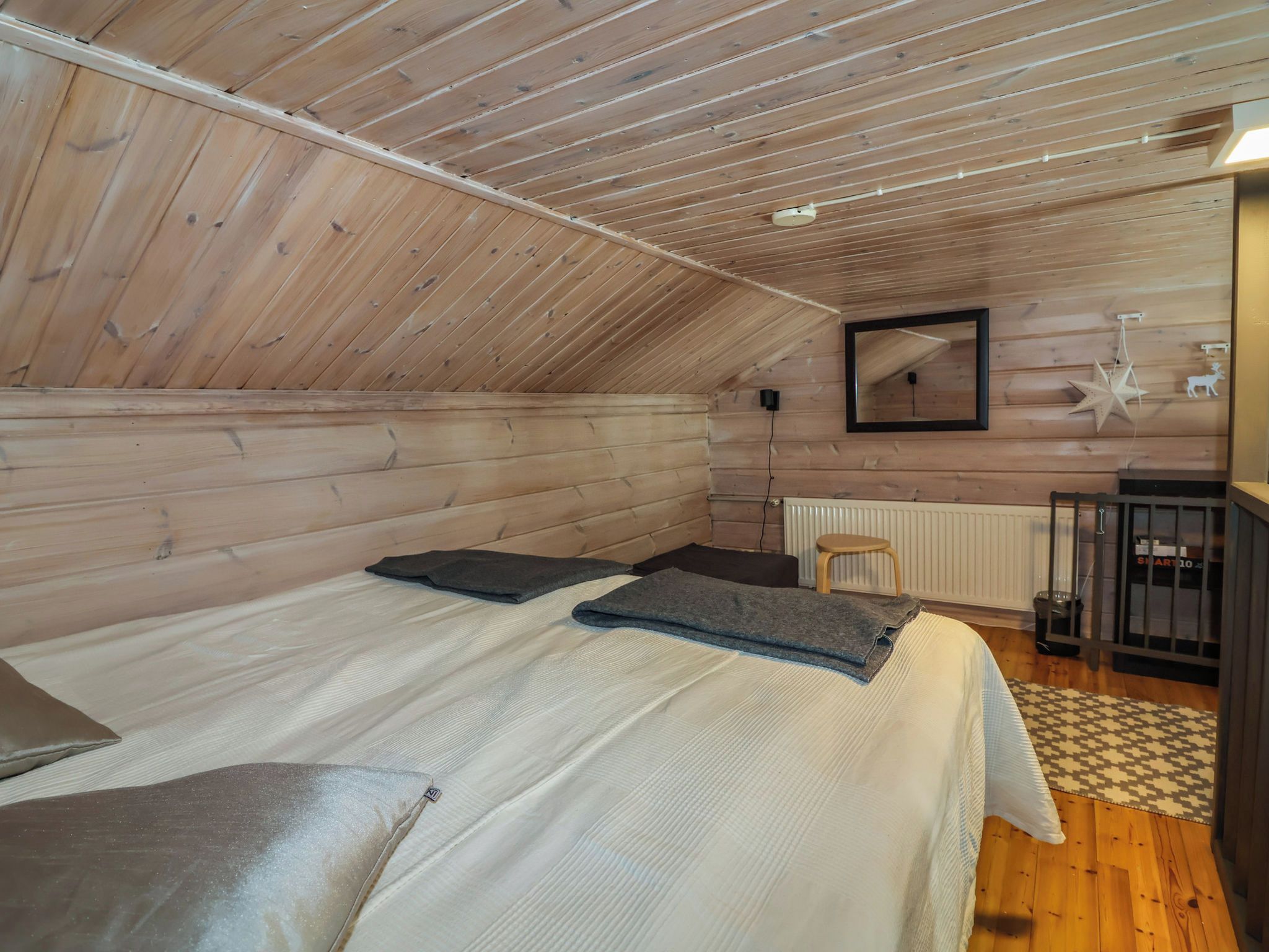 Une pièce avec un plafond en pente, des murs en bois, un lit avec des oreillers, un miroir, un radiateur et un petit tabouret.