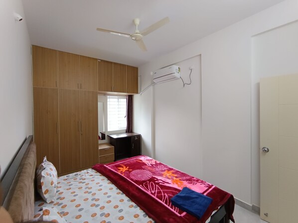 Een slaapkamer met een bed, een kledingkast, een plafondventilator, een airconditioning en een raam met gordijnen.