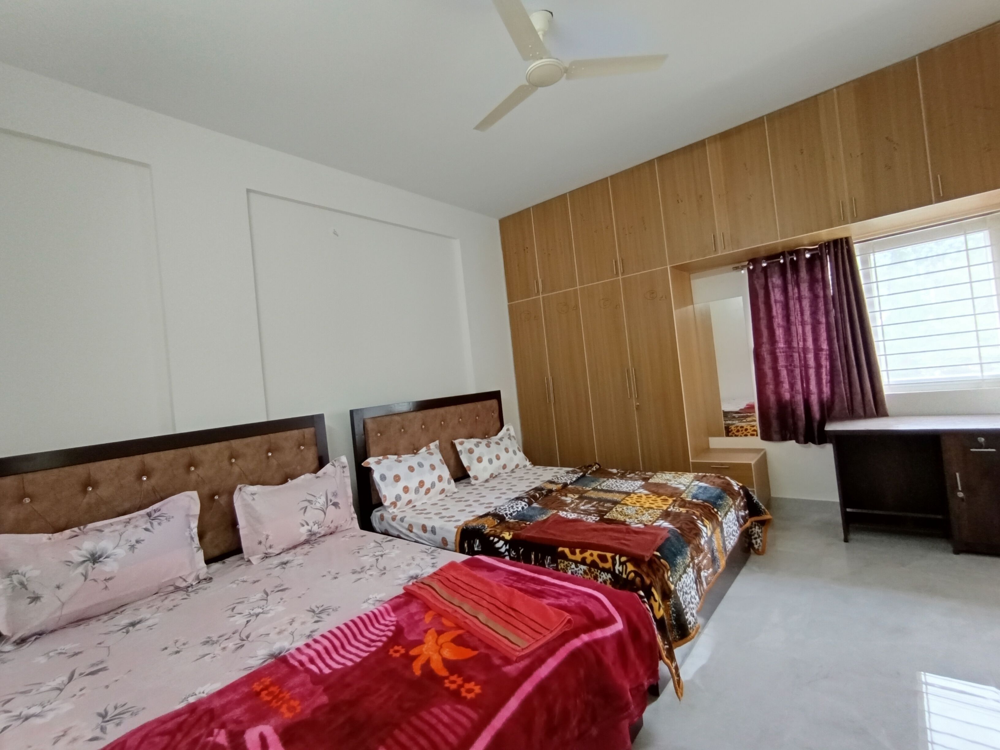 A bedroom with two beds, wooden wardrobe, ceiling fan, and a window with curtains.