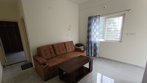 A spacious living room with a brown sofa, a wooden coffee table, and a window with curtains. - SAGAR NIWAS SARJAPUR (Bengaluru)