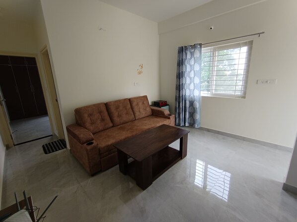 A spacious living room with a brown sofa, a wooden coffee table, and a window with curtains. - SAGAR NIWAS SARJAPUR (Bengaluru)