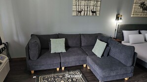 Un salon moderne avec un canapé d’angle gris foncé, deux coussins de couleur vert clair, un tapis à motifs noir et blanc et un lit avec une literie blanche.