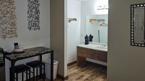 A compact kitchenette with a microwave, a small table, and two stools. Adjacent to the kitchenette is a bathroom with a sink, mirror, and wooden floor. - Modern in the Mountains | Free Shuttle to SDC (Branson)