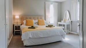 Une chambre à coucher bien rangée, avec un lit, des tables de chevet, une coiffeuse avec un miroir et une fenêtre avec des rideaux.