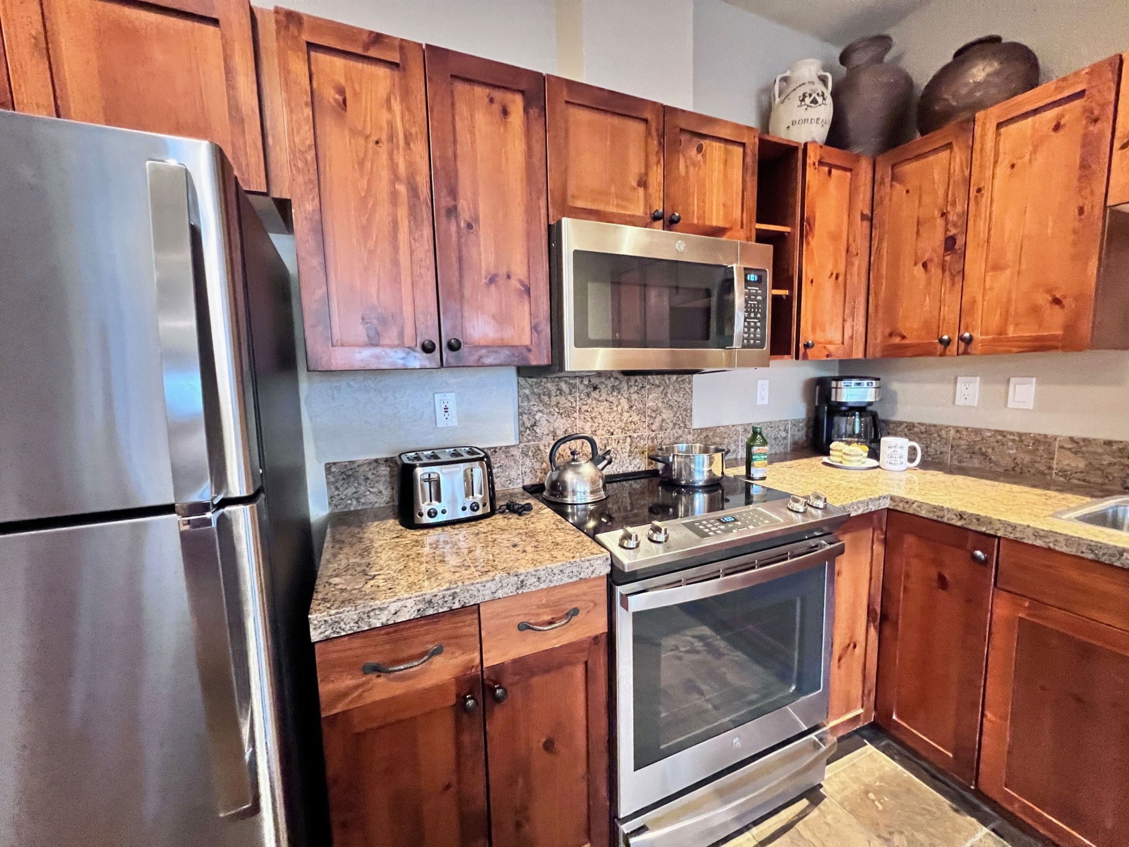 A kitchen with wooden cabinets, a stainless steel refrigerator, a microwave, a toaster, a kettle, a coffee maker, and a stove.