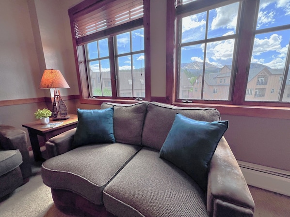 A living room with a sofa, a lamp, and a view of buildings through large windows.