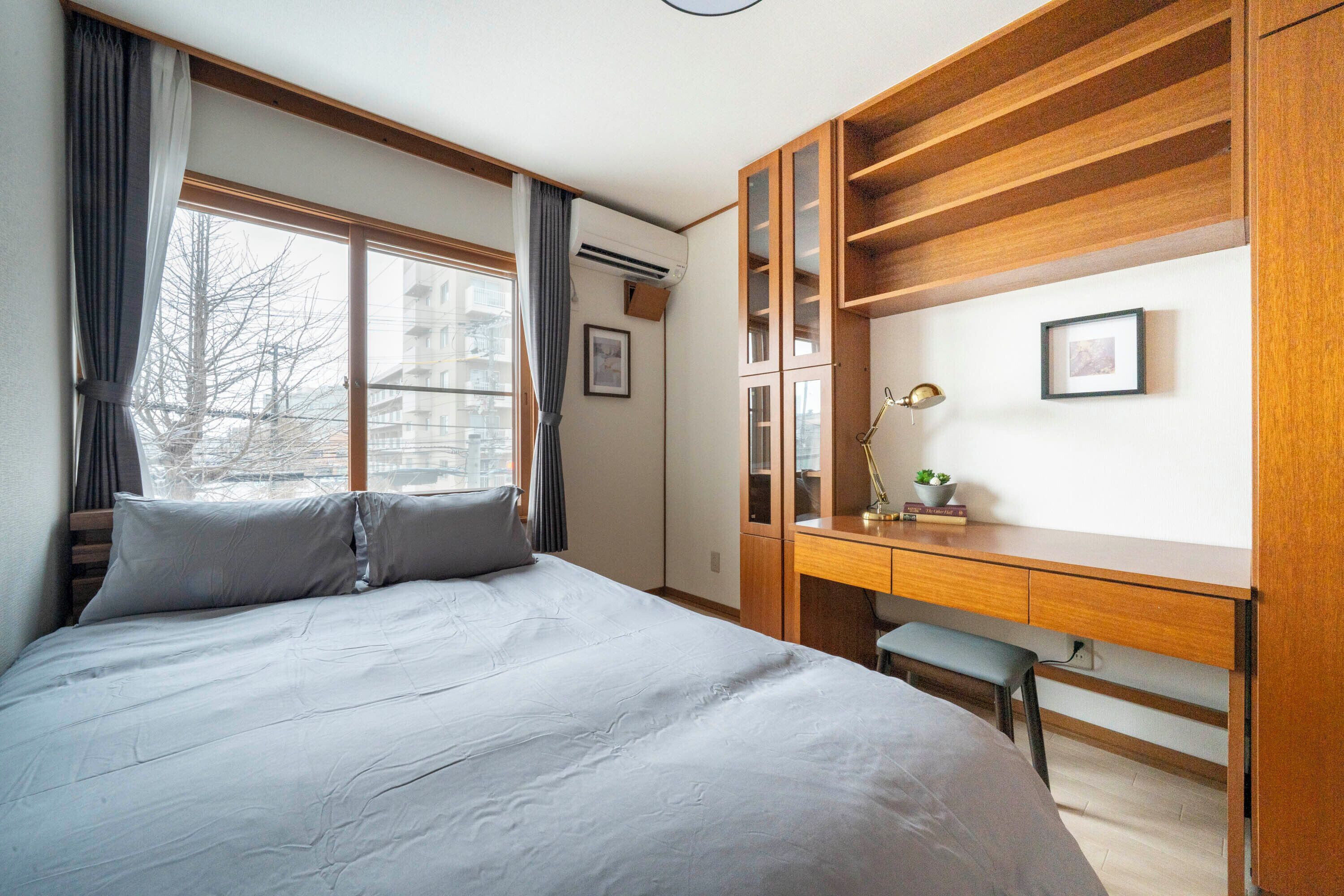 A bedroom with a bed, a desk, and a chair. There is a window with curtains, an air conditioner, and a framed picture on the wall.
