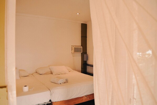 A single bed with a white mattress and pillows, a wall-mounted air conditioner, and a roll of toilet paper on the floor.