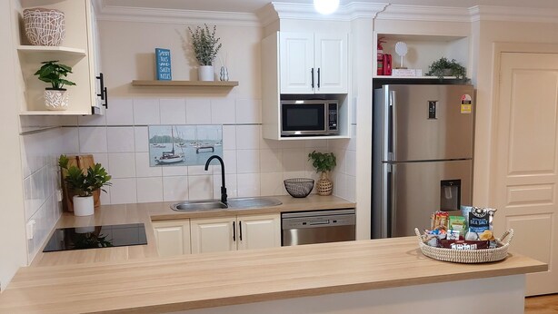 A modern kitchen with white cabinetry, a stainless steel refrigerator, a microwave, and a sink. There are potted plants and a decorative bowl on the shelves.