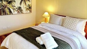 A bedroom with a bed, a ceiling fan, a painting of palm trees, and a lamp.