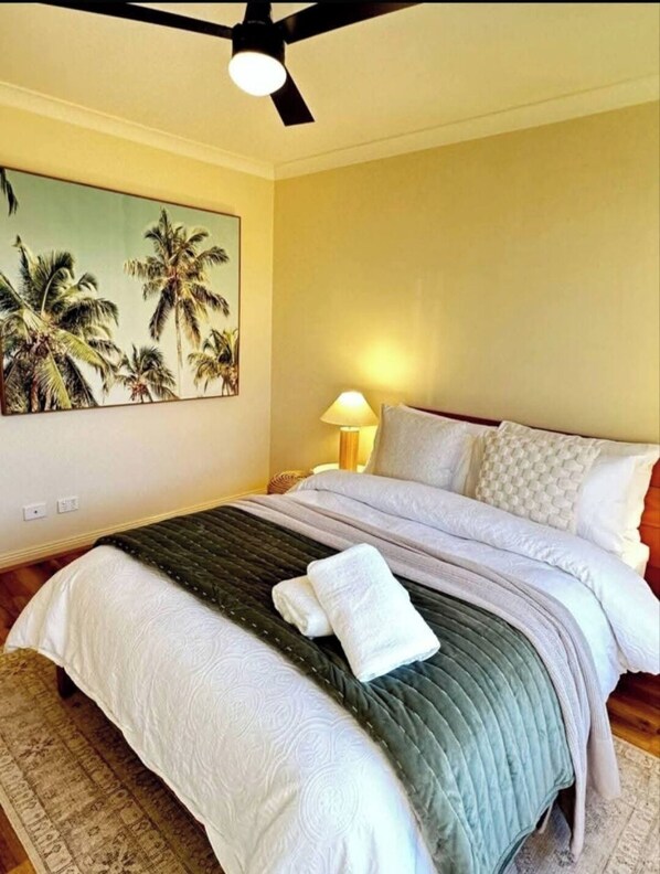 A bedroom with a bed, a ceiling fan, a painting of palm trees, and a lamp.