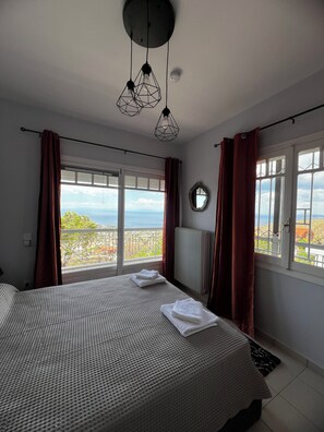 A bedroom with a large bed, two hanging pendant lights, a view of the sea, and a window with curtains. - Panorama  Escape  Villa (Saronida)