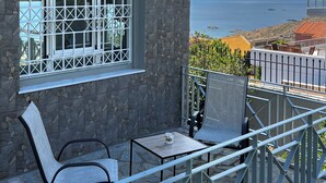 Ein Balkon mit Tisch und Stühlen, ein Fenster mit Metallgitter und Blick auf das Meer und die fernen Hügel.