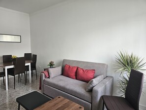 A living room with a grey sofa, red cushions, a wooden coffee table, a dining table with chairs, and a potted plant.