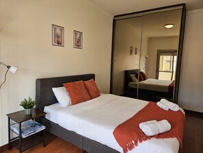 A neatly made bed with white and orange bedding, a small bedside table with a lamp, and a mirror reflecting the room.
