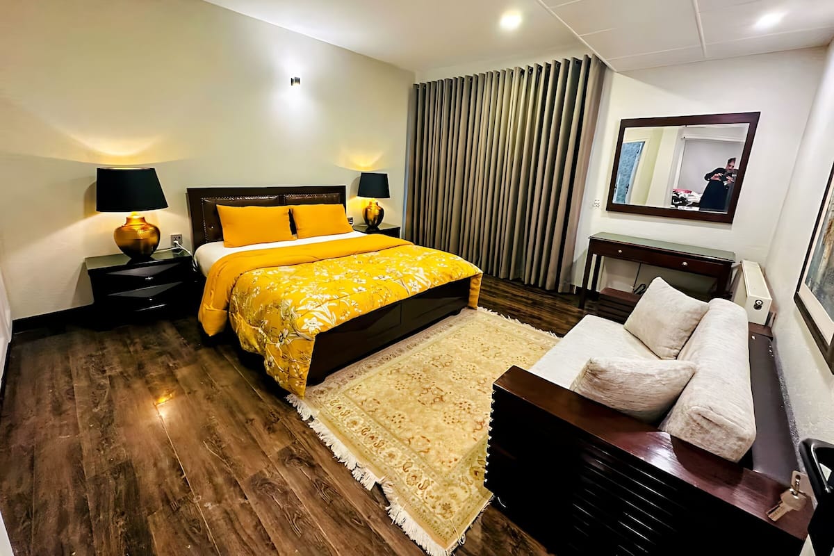 Habitación de hotel con una cama grande, una mesita de noche con lámparas, suelo de madera, alfombra, un sofá y un espejo en la pared.