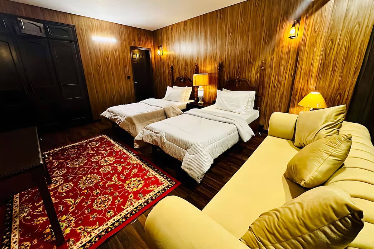 Habitación de hotel con dos camas, un sofá, un tapete rojo estampado, paneles de madera y lámparas fijadas a la pared.