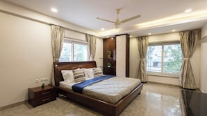 A bedroom with a wooden bed, a nightstand, a ceiling fan, and large windows with curtains. - Victoria Apartment by Jadecaps (Kolkata)
