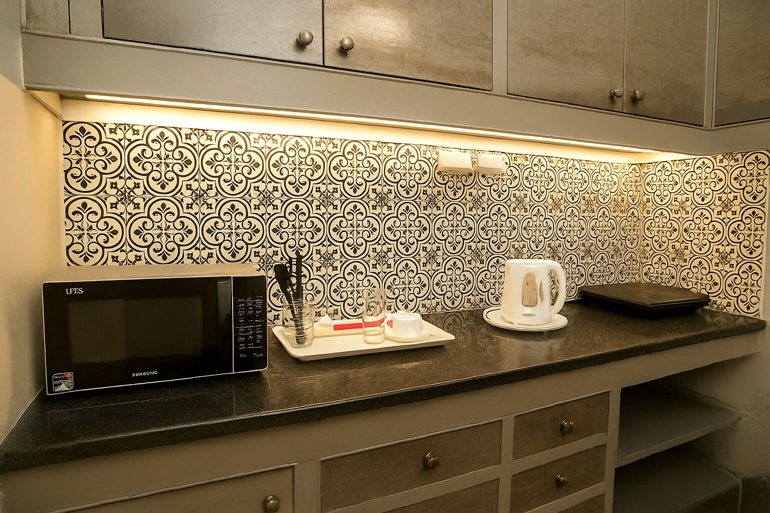 Una cocina con microondas, una bandeja con utensilios, una tetera blanca y un salpicadero decorativo.