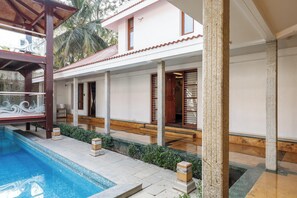 A poolside area with a pool, a covered seating area, and a building with a tiled roof and wooden doors. - Ananta Vilas by Stayjade (Bengaluru)