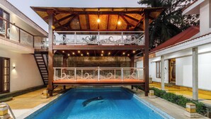 A swimming pool with a dolphin, a covered deck area, and a staircase leading to a second floor. - Ananta Vilas by Stayjade (Bengaluru)